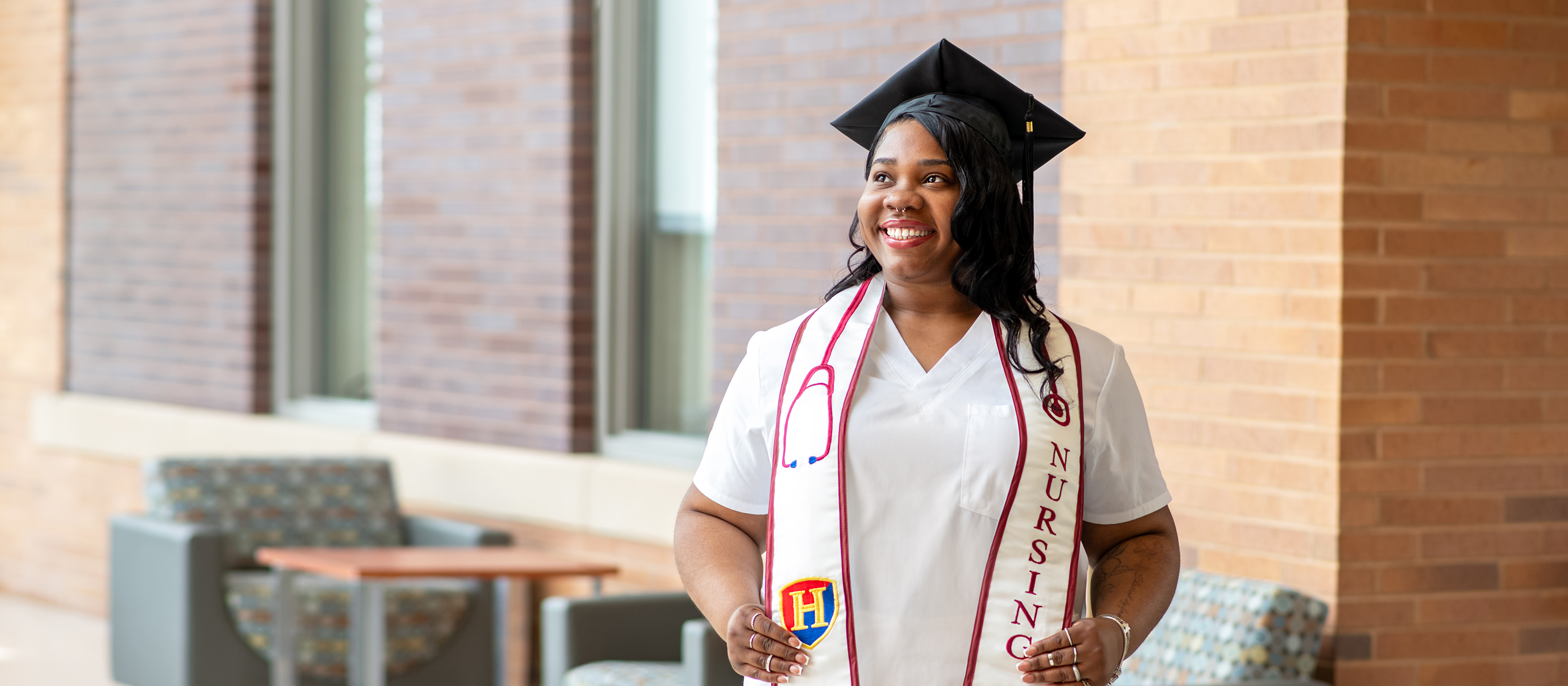 A woman in a white nursing uniform and graduation cap stands indoors, smiling. She holds a diploma and wears a stethoscope and a 
