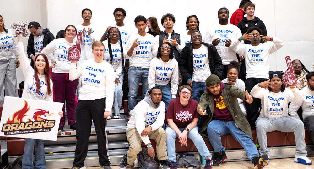 A diverse group of college students wearing matching “Follow the Leader” shirts pose together on indoor bleachers, smiling and making hand gestures, with one student holding a “Dragons – Howard Community College” sign and others holding foam fingers.