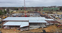 A wide, aerial view of an active construction site featuring a large steel-frame building in progress. Metal decking covers portions of the structure, while cranes, construction vehicles, and workers are visible around the site. Trees, roads, and additional buildings appear in the background under an overcast sky.