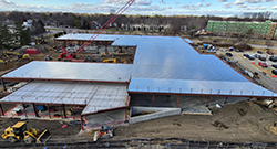 A wide, aerial view of an active construction site featuring a large steel-frame building in progress. Metal decking covers portions of the structure, while cranes, construction vehicles, and workers are visible around the site. Trees, roads, and additional buildings appear in the background under an overcast sky.