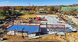 Aerial view of building construction site with steel framing, concrete foundation, and crane amid fall trees.