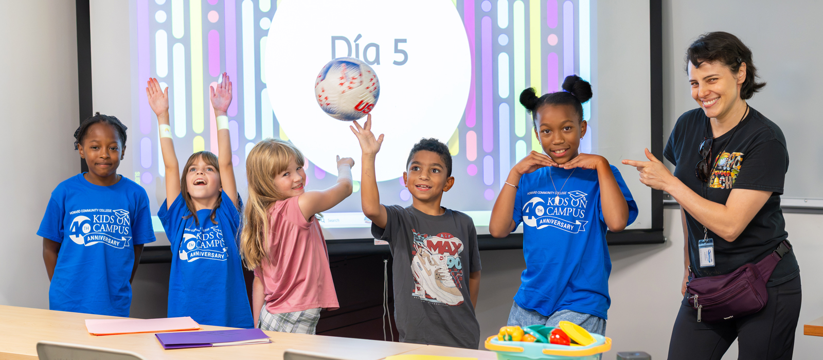 Five children participate in a Kids on Campus activity in a classroom, with one child tossing a soccer ball while an instructor points toward the group. A presentation screen and colorful activity materials are visible in the background.