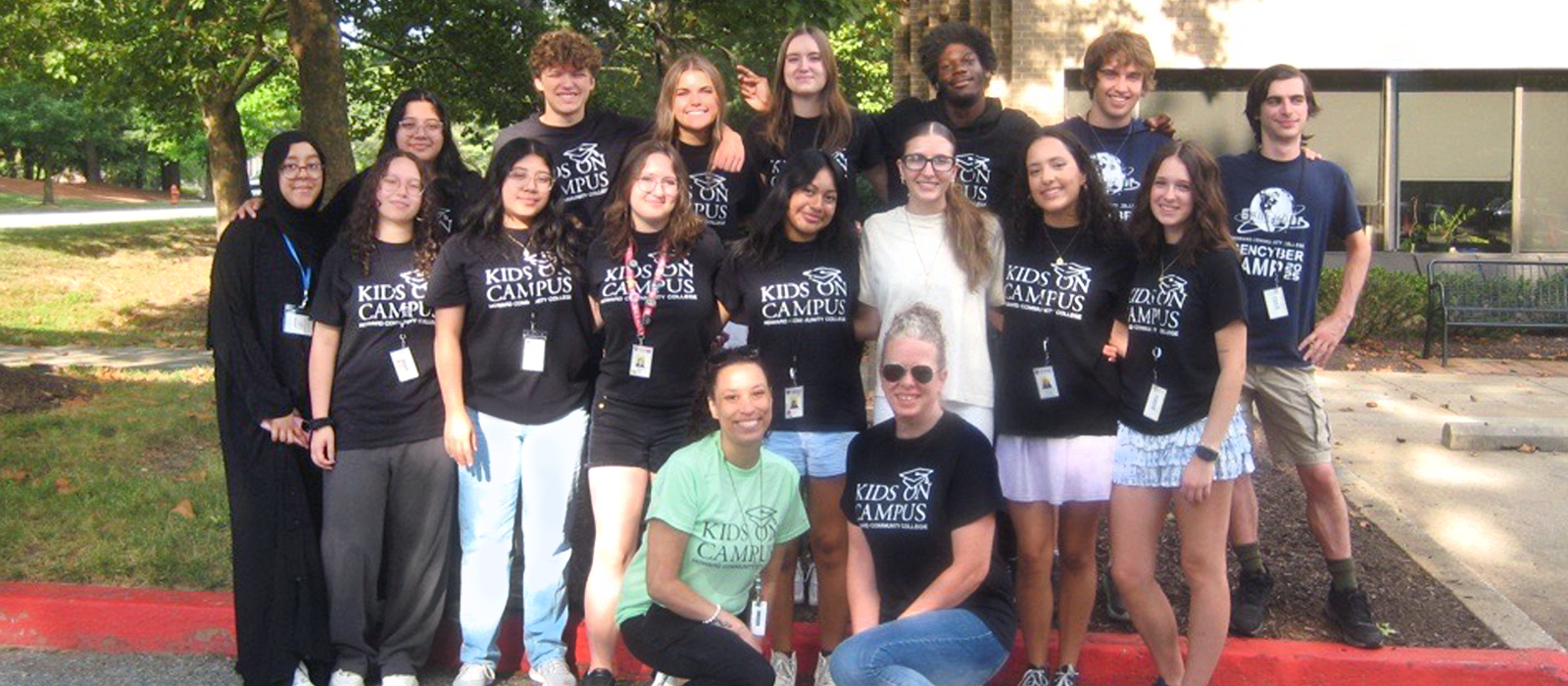 Group of smiling Kids on Campus staff and aides posing outdoors in front of a college building, wearing matching program T-shirts.