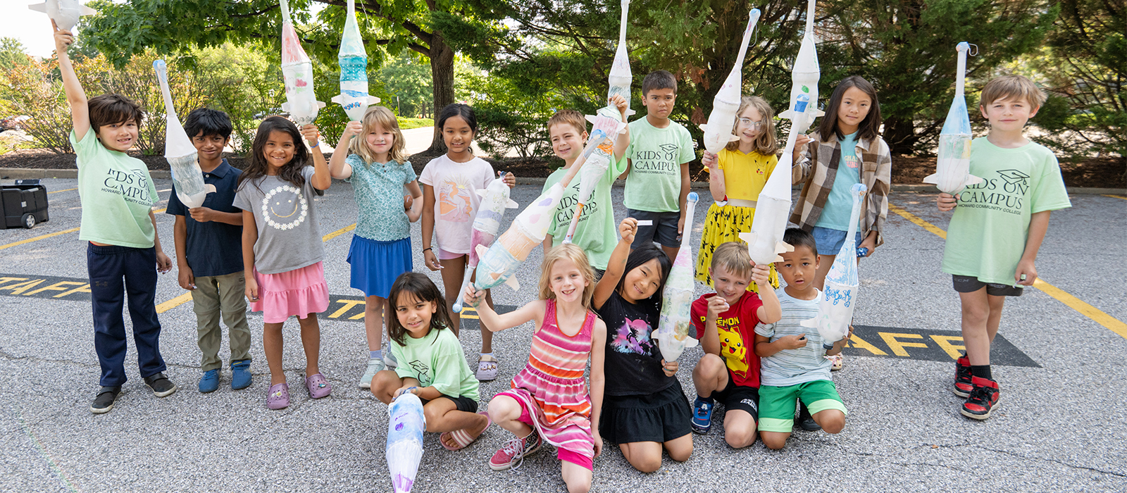 Group of smiling children at Howard Community College’s Kids on Campus program holding up handmade rocket crafts outdoors.
