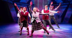 Six energetic performers in school uniforms strike dynamic dance poses on a brightly lit stage, surrounded by colorful, geometric set pieces and vertical light bars, evoking the vibrant atmosphere of a theatrical musical performance.