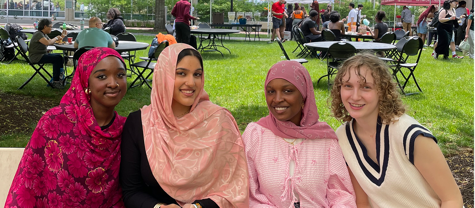 Four students sit together and smile during an outdoor campus event with picnic tables and people in the background.